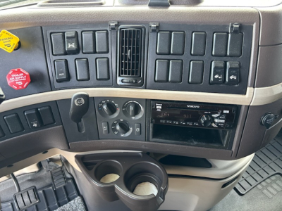 Cab interior of a Volvo truck dashboard showing multiple blank switch panels, central air vents, Volvo stereo, climate dials, cup holders with items, and a gear shifter at left.
