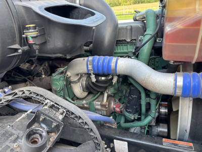 Engine bay with turbo piping and blue silicone hoses visible in a Volvo VNL780. Green engine block, black intake, and various hoses and belts.