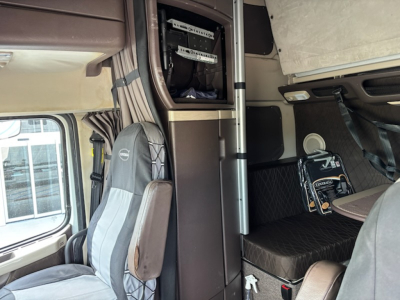 Truck cab interior with storage cabinet and sleeper area; Volvos branding visible on seat; small label panels and a toolbox or electronics box mounted behind the driver seat.