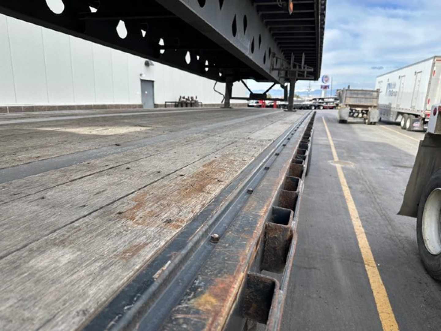 Flatbed trailer edge with steel deck showing wear and rust-like stains and railings; long side rail and wheel area visible under a parked trailer