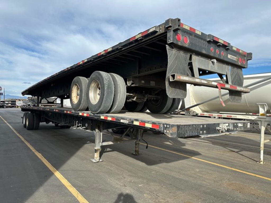 Manac steel flatbed trailer in raised position showing tandem axles and rear overhang with reflective tape and mounting hardware.