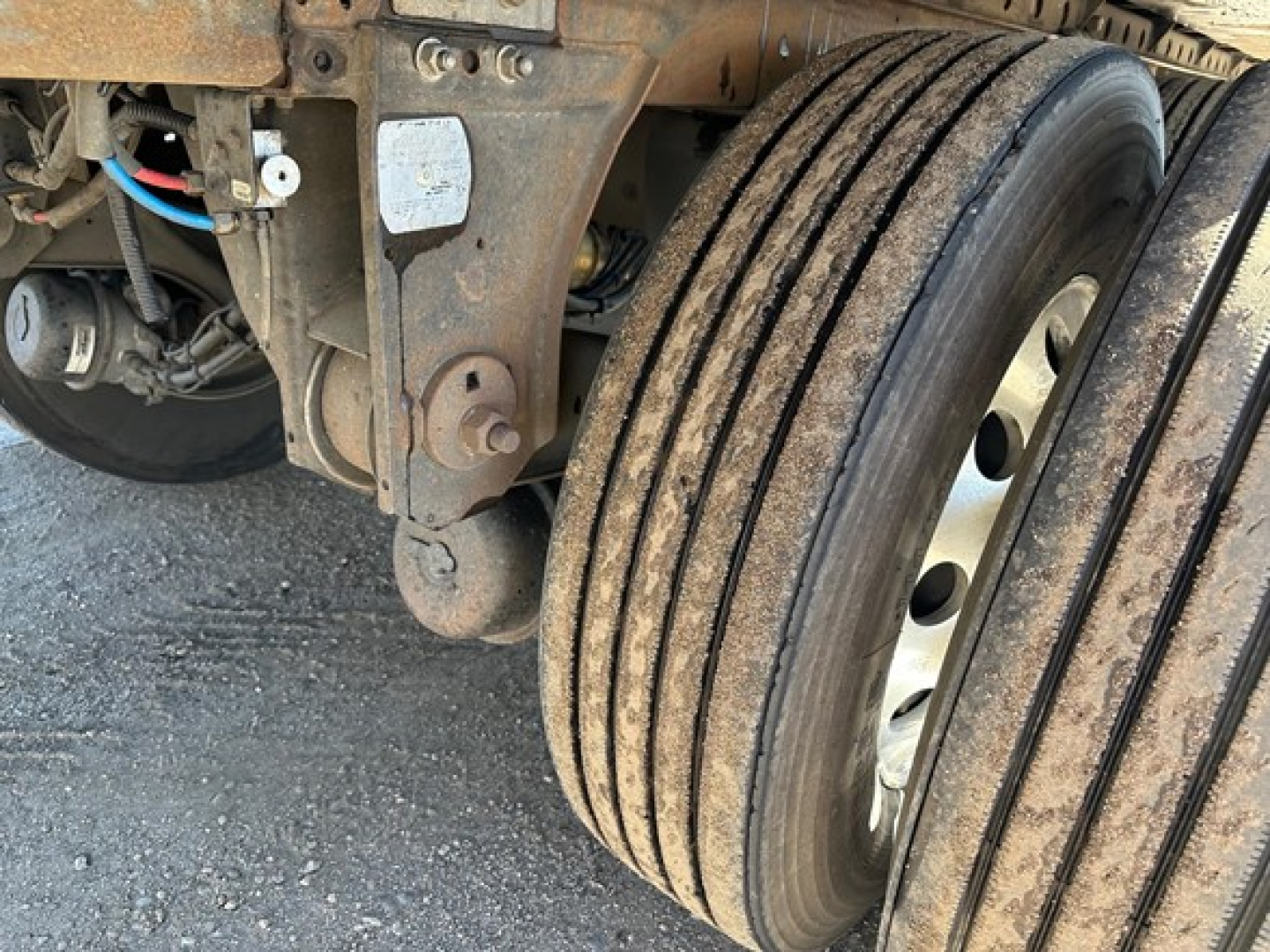 Trailer dual-axle wheel assembly with visible suspension components and electrical wiring; rusted metal frame and mud-coated tires.