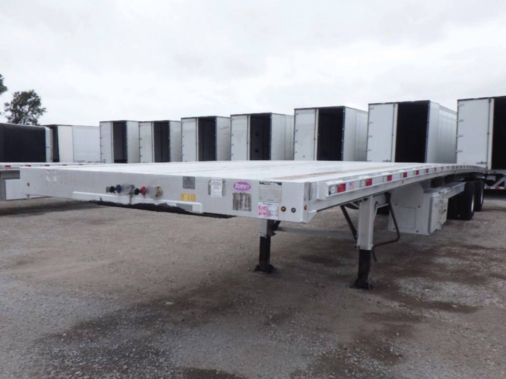 Flatbed trailer showing aluminum deck with landing gear and wheel wells in a lot.