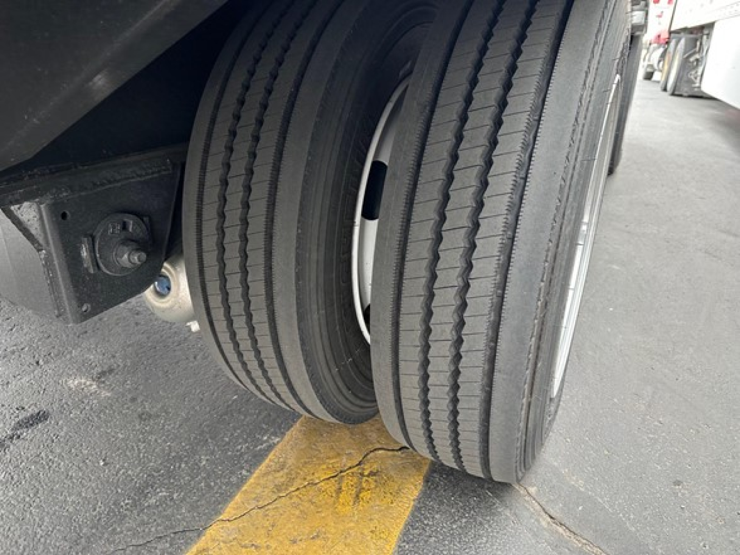 Dual rear trailer tires showing tread and wheel hub with part of the chassis and yellow painted line on pavement.