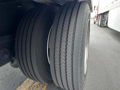 Dual rear trailer tires in view with ribbed tread pattern and metal wheel rims under a drop-deck trailer.