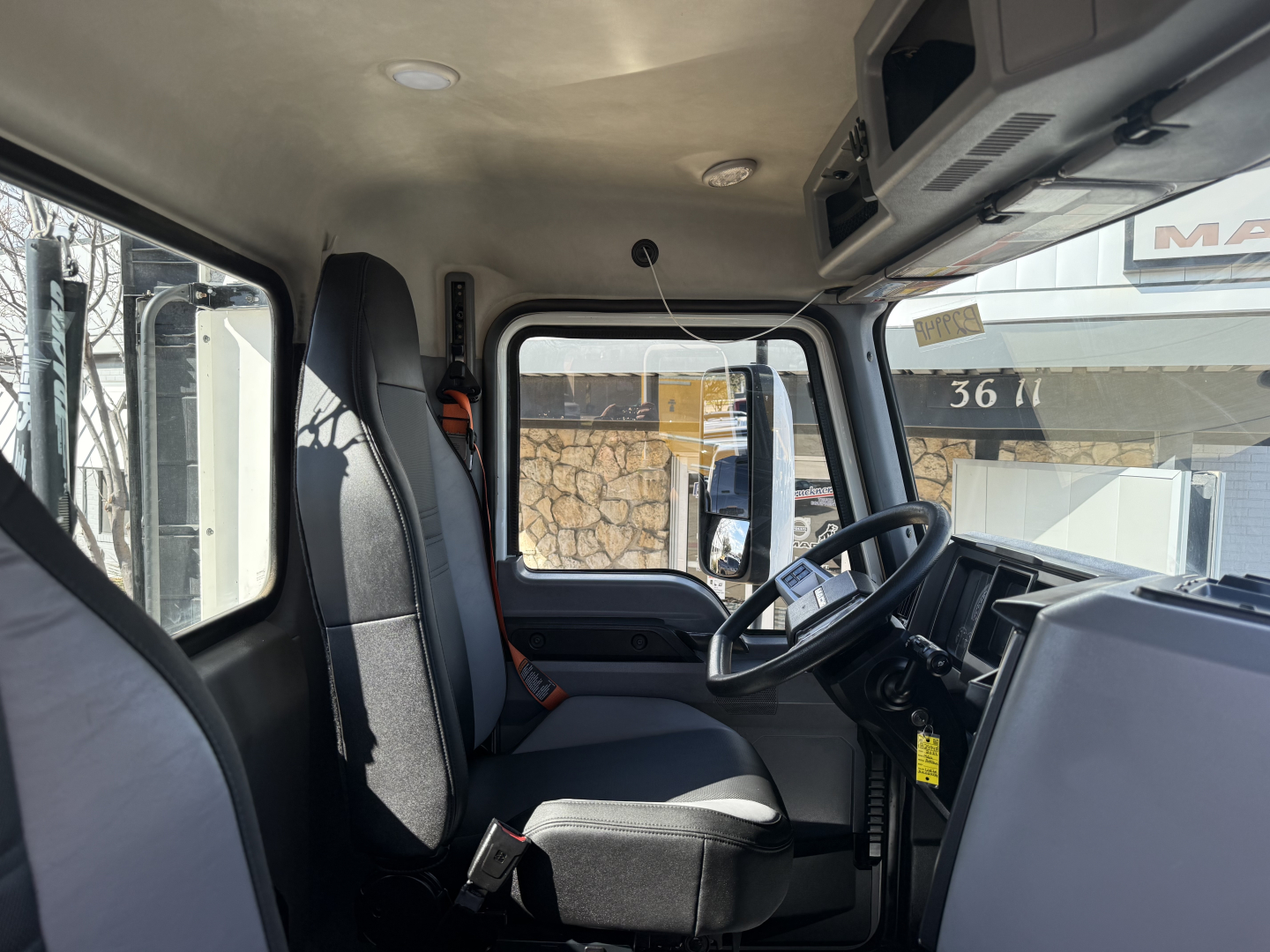 Truck cab interior of 2022 Mack AN42T showing driver seat, steering wheel, dashboard, and front window with overhead storage cabinet.