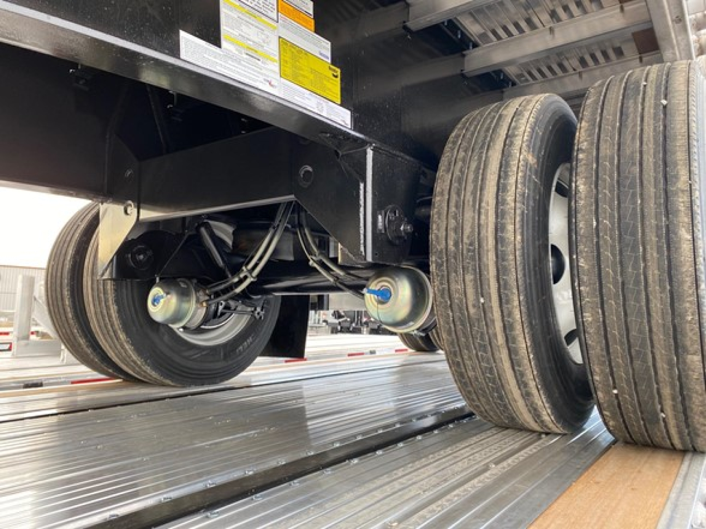 Trailer axle and wheels under a flatbed trailer showing dual rear tires and undercarriage components.
