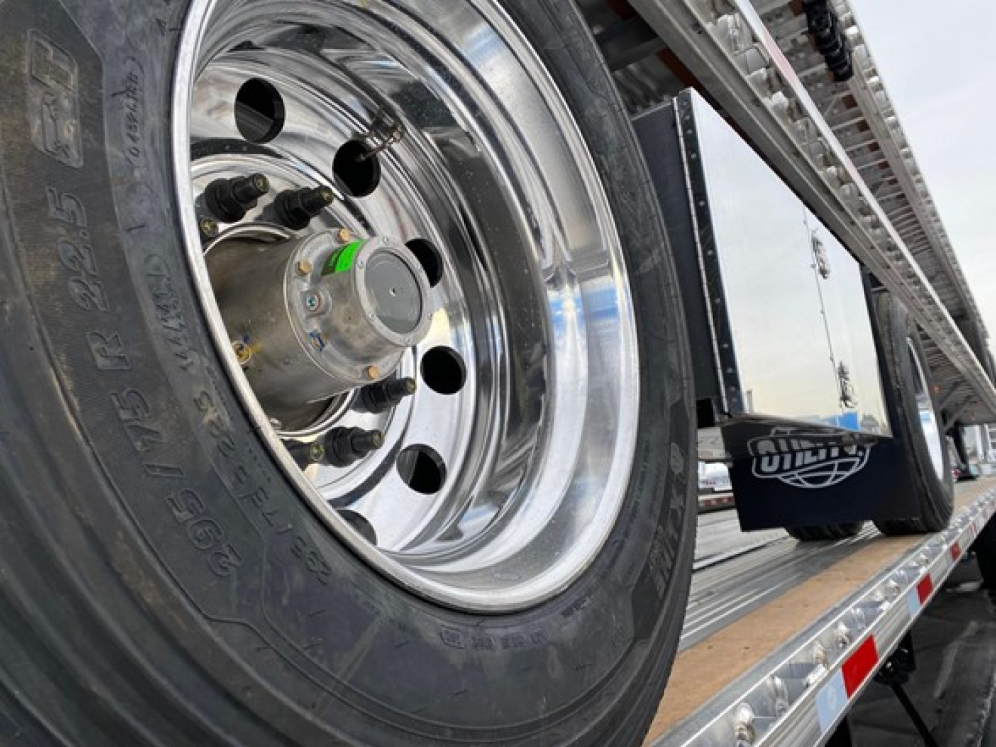 Close-up view of a shiny chrome wheel on a flatbed trailer with a green valve cap and lug nuts; part of a long metal trailer deck with reflective strips.