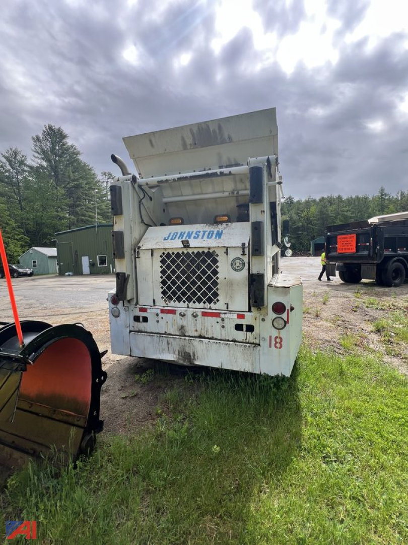 Rear view of a Johnston sweeper with a mesh grille, large white body, and blue Johnston branding; equipment shows a hopper and tailgate area.