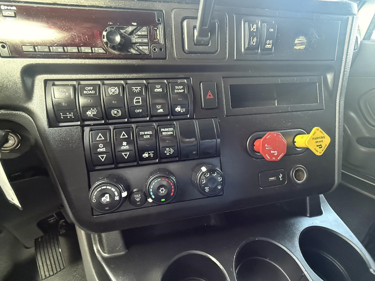 Truck cab dashboard showing controls and gauges in a 2020 International LT sleeper. Visible switches, hazard button, climate knobs, and circular red and yellow knobs; partial radio slot.