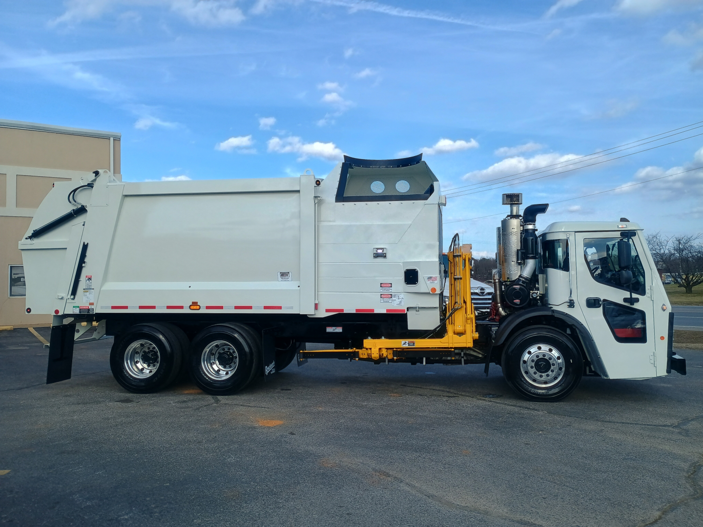 2026 Mack LR64 truck side view with refuse body and hydraulic lifting mechanism, white cab and container, multiple wheels visible.