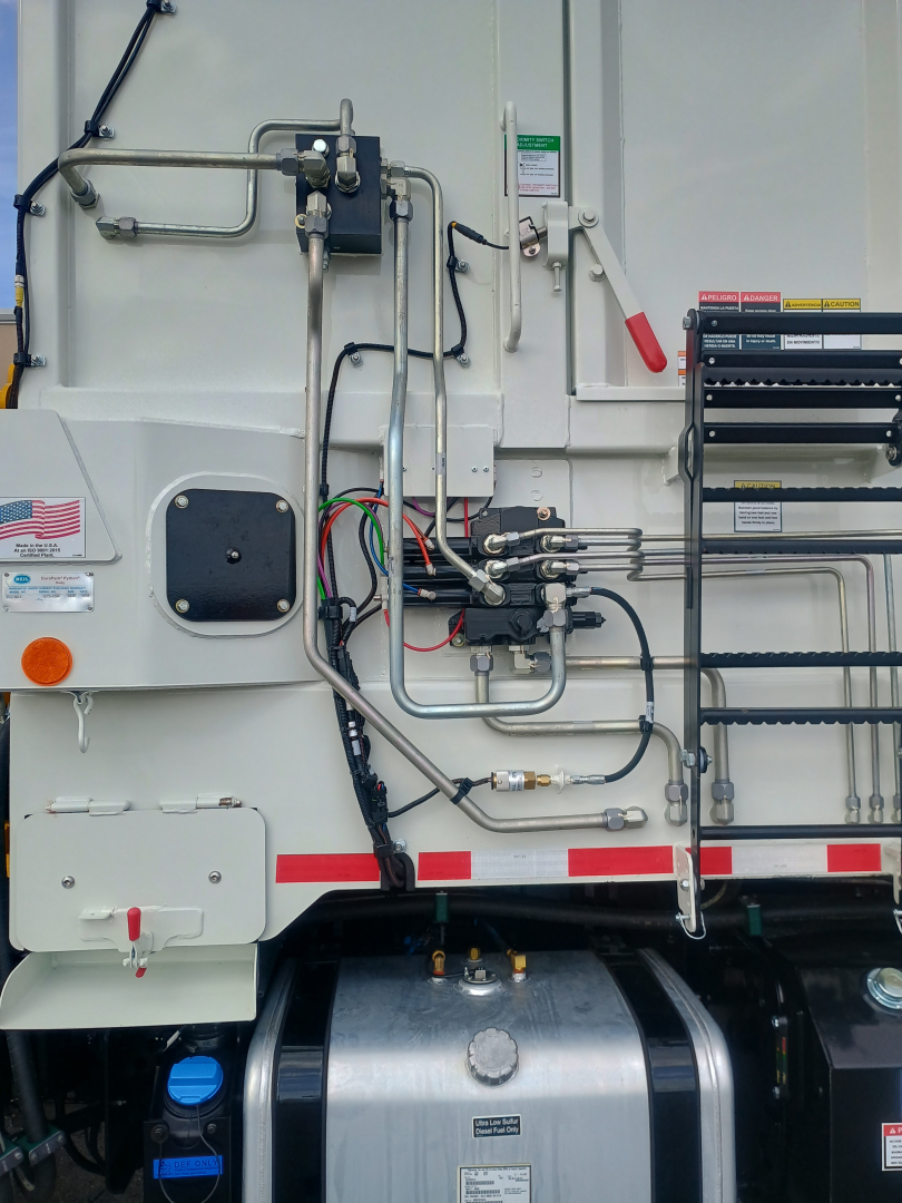 Side of a 2026 Mack LR64 refuse truck showing hydraulic lines, control valves, and metal piping on white exterior.