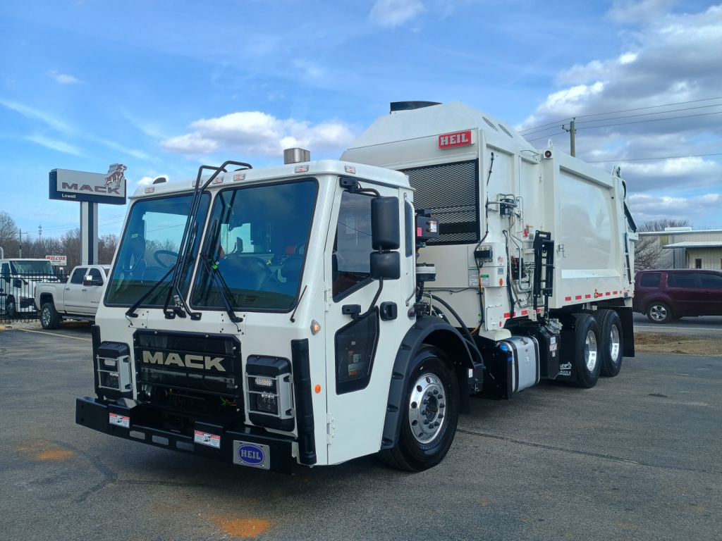 Truck cab and refuse body of a 2026 Mack LR64 in dealership lot, side loader configuration with white cab and dual rear axles; visible Mack logo on grille and HEIL branding on refuse body.