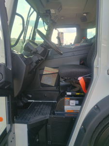 Truck cab interior of a 2026 Mack LR64 side loader showing steering wheel, driver seat, dashboard, and entry area; visible labels and safety stickers in the footwell.