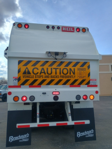 Rear view of a white Mack LR64 refuse truck showing caution sign panel, lights, and mud flaps with Bruckners Truck & Equipment logos.