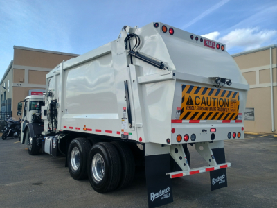 New Mack LR64 refuse truck in showroom lot with caution decal on rear, dual rear axles, white body, rear lights, and black Buckner’s mud flaps.