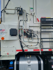 Side of a 2026 Mack LR64 refuse truck showing hydraulic lines, control valves, and metal piping on white exterior.