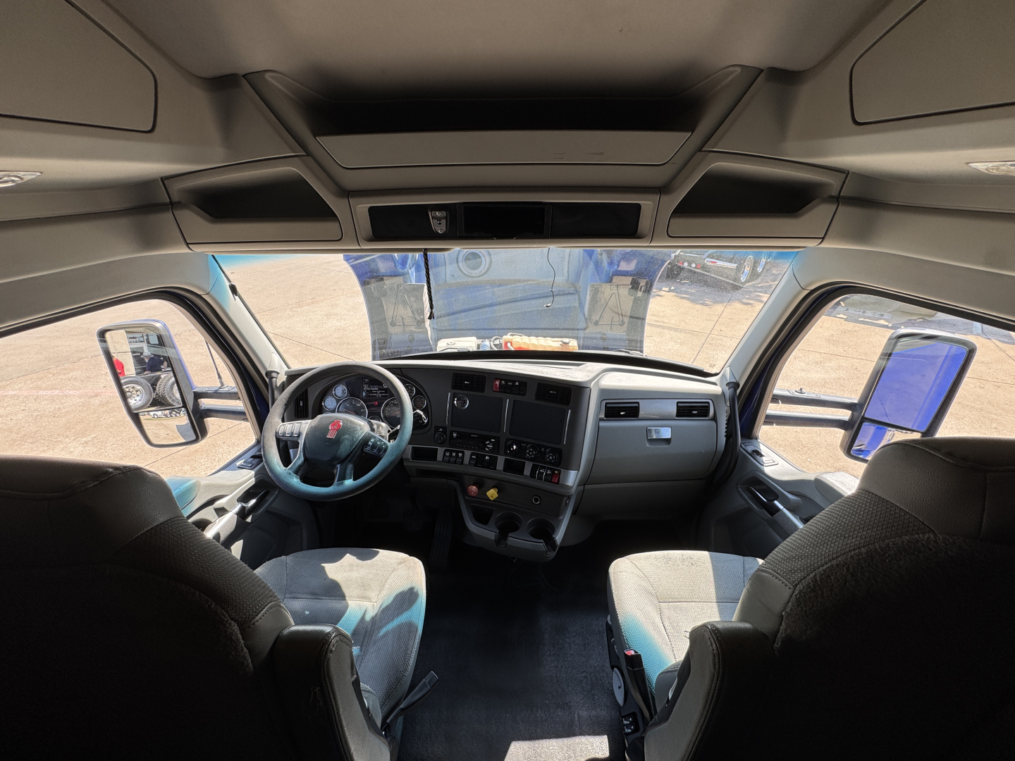 Kenworth T680 cab interior showing steering wheel, dashboard, and front seats from passenger side, with windshield and mirrors visible.