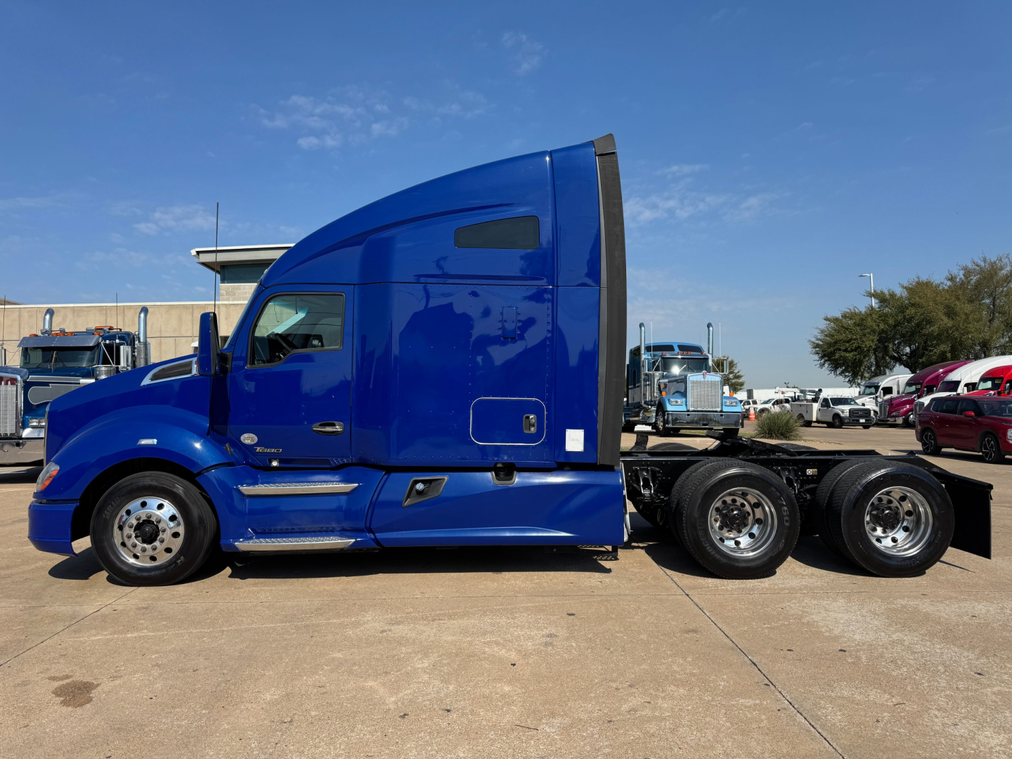 Kenworth T680 side view in blue cab with sleeper, 2019 model, 6x4 tractor chassis, dual rear axles, gravel lot, clear sky.