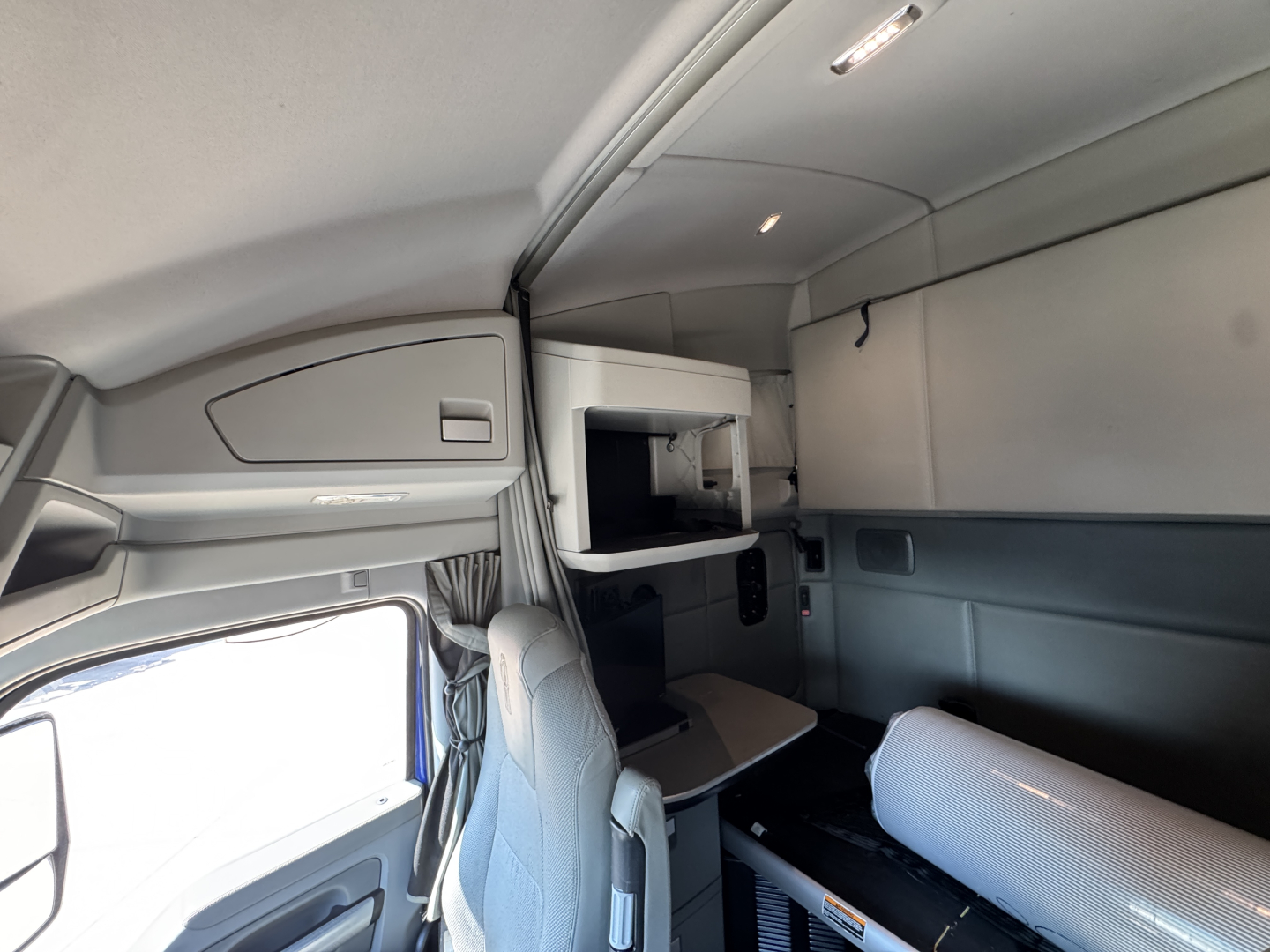 Kenworth T680 sleeper cab interior showing upper bunk area, storage cabinet, and seating with light gray upholstery.