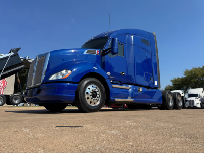 Blue Kenworth T680 tractor in-profile view showing front fender, hood, and cab with sleeper section; chrome grille, large side mirrors, and front wheel; clear sky background.