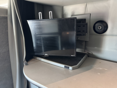 In-vehicle TV monitor mounted in a Kenworth T680 sleeper cab with a curved desk surface and speaker panel, showing a small flat-screen monitor and mounting hardware.
