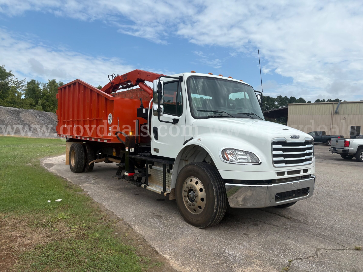 Truck cab of a 2023 Freightliner M2 106 with a large orange boom/machine on the back, side view showing front grille and wheel hubcaps.