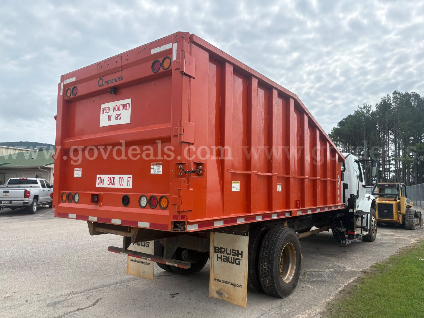 Freightliner M2 106 dump body rear view showing Bright red container with multiple labels and lights; small white license plate area; mud flaps reading BRUSH HAWG; rear bumper and taillights visible.