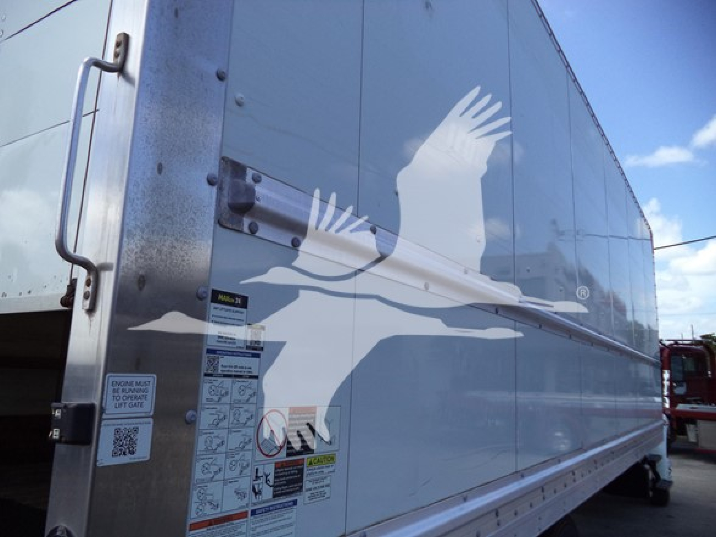 Truck box side with various safety and instruction labels near a door; cargo trailer style with riveted panels and a white logo overlay image of a bird in flight.