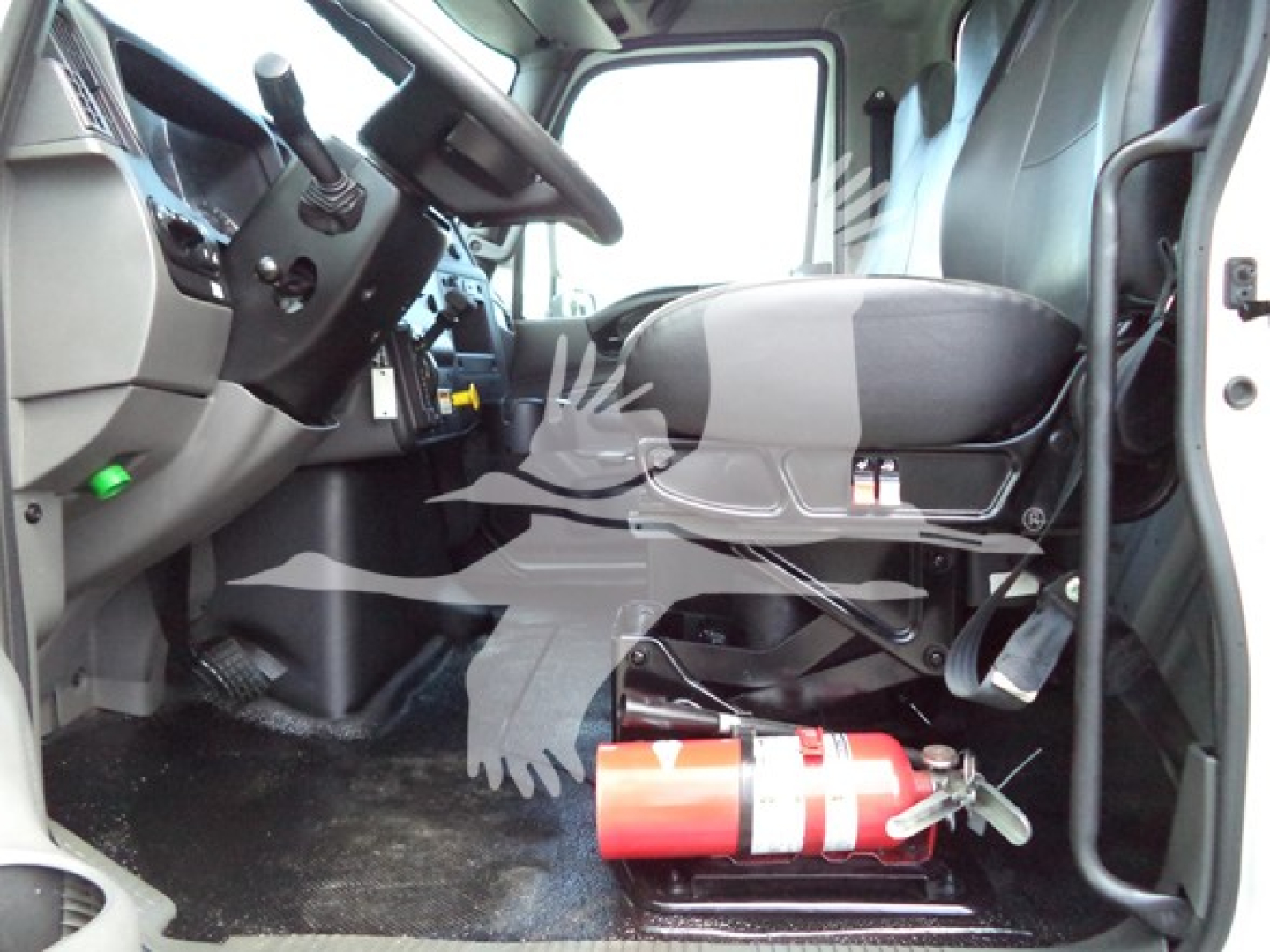 Cab interior of a 2021 International MV truck showing driver seat, steering wheel, dashboard, pedals, and a mounted red fire extinguisher under the seat.