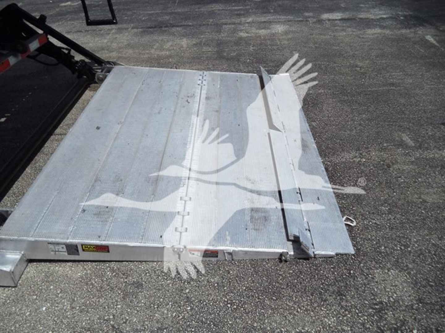 Truck bed area showing aluminum flatbed with folding ramp in metal panels and hinge joints, open ground surface visible, light reflections on metal.