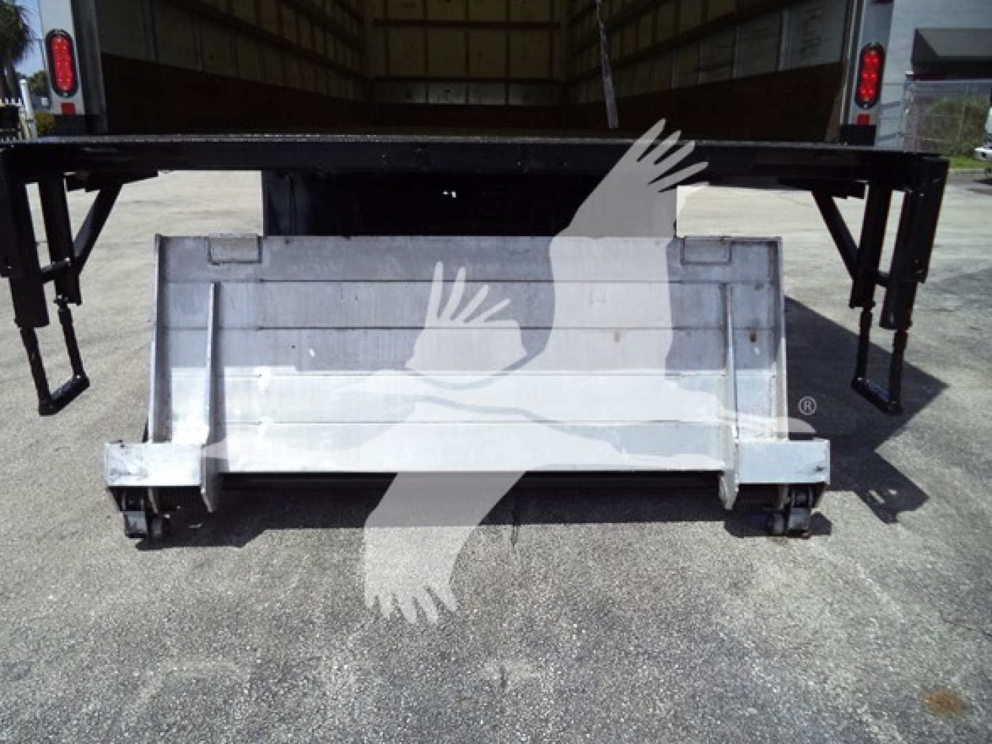 Truck rear view showing a metal cargo bed or bumper with lifting legs and wheels on a trailer. Clear text not visible.