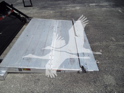 Truck bed area showing aluminum flatbed with folding ramp in metal panels and hinge joints, open ground surface visible, light reflections on metal.
