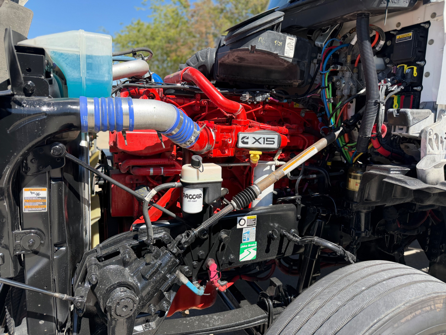 Kenworth T880 engine bay view showing 6-cylinder red engine with 6XIS badge, blue intercooler hose, hydraulic lines, and various wiring harnesses.