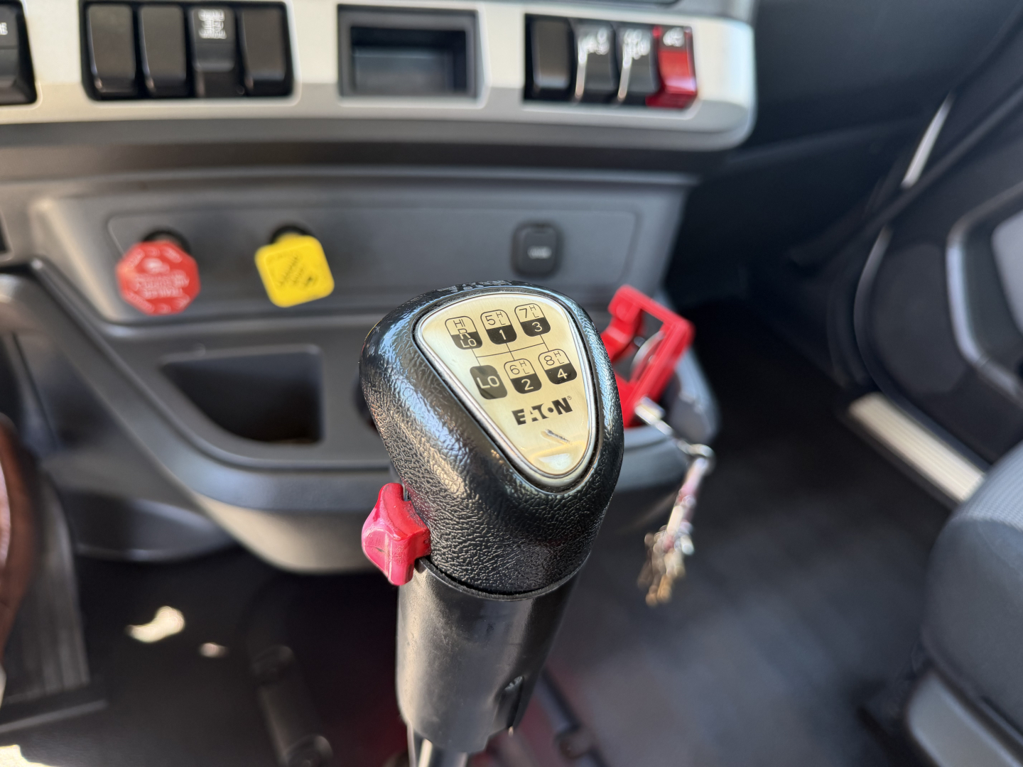Kenworth truck interior showing gear selector with labeled shift pattern and red controls; dashboard with controls and ignition area visible.