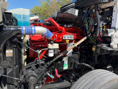 Kenworth T880 engine bay view showing 6-cylinder red engine with 6XIS badge, blue intercooler hose, hydraulic lines, and various wiring harnesses.