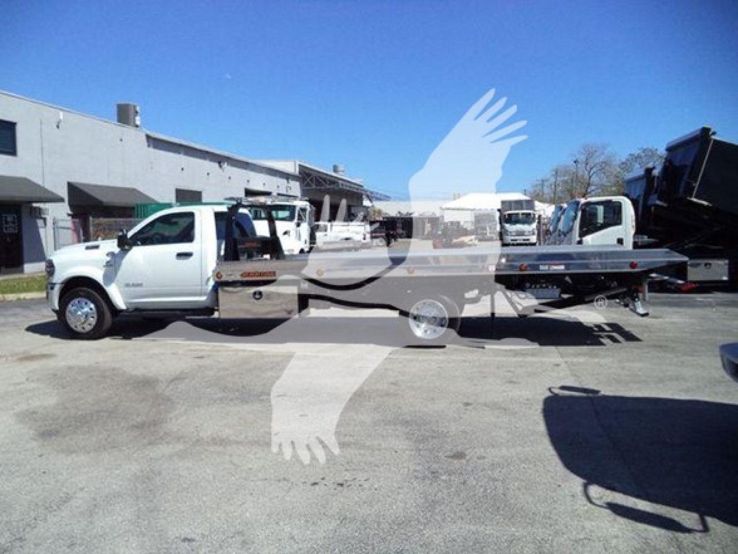 Truck with tow truck bed in a lot, Ram 2026 Big Horn new vehicle, side view showing cab and flatbed with equipment in background.