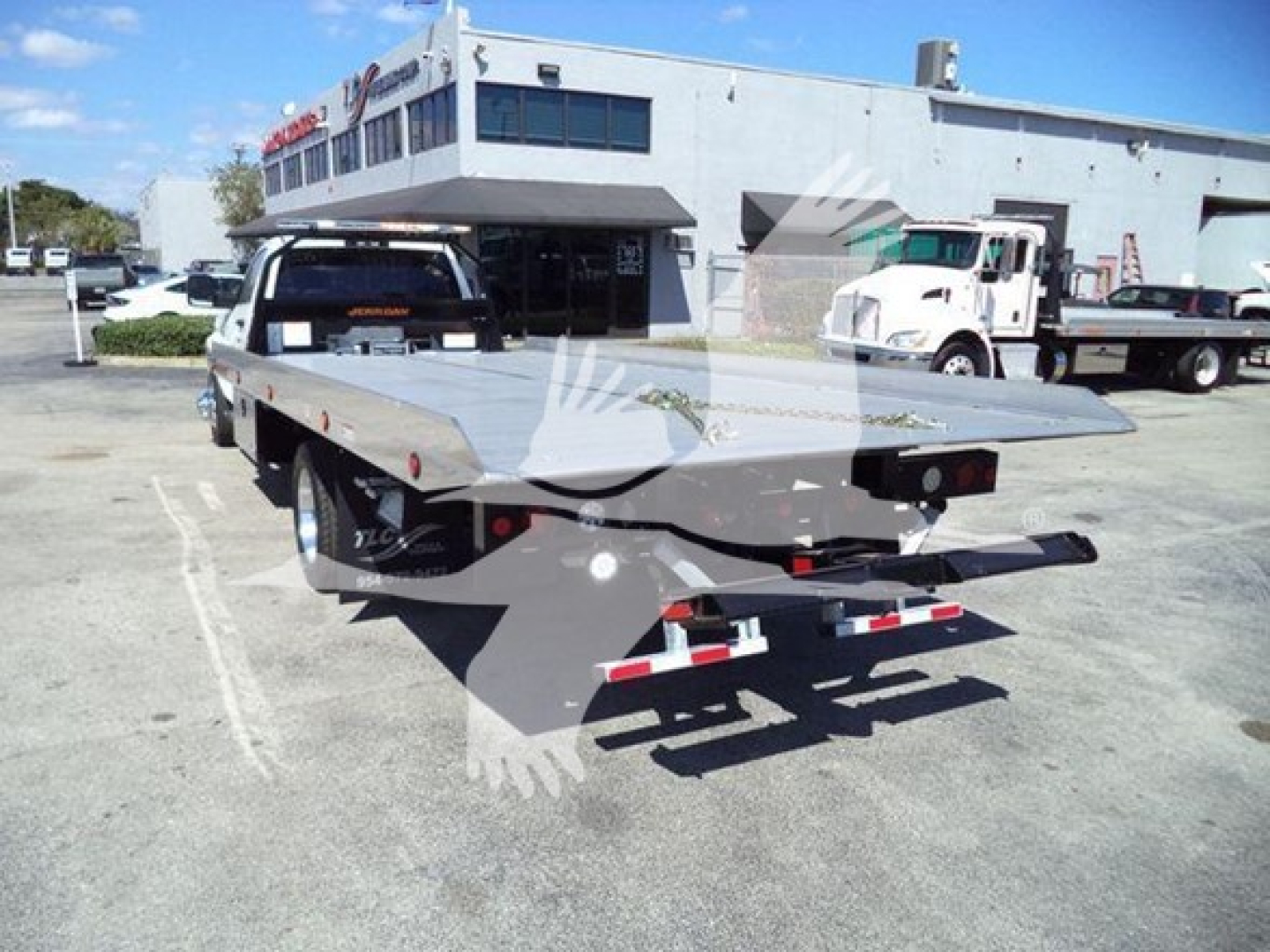 Flatbed rollback truck trailer in a dealership lot, viewed from rear left side. Visible text on the trailer accents and taillights; background shows a white building and other trucks.