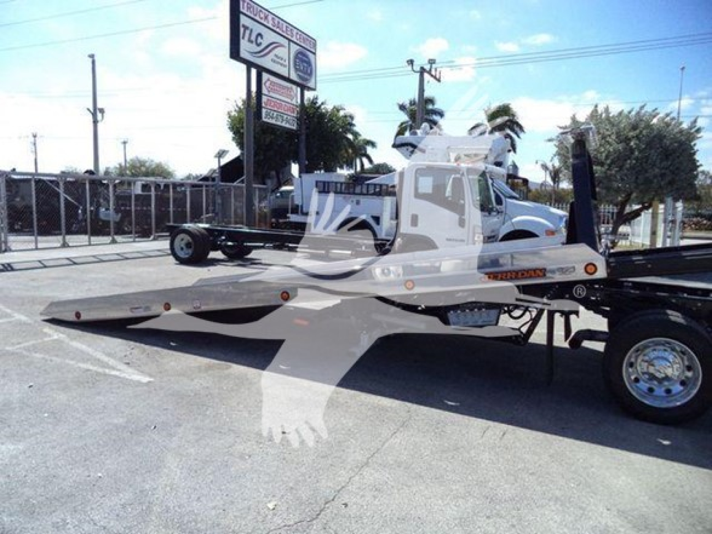 Roll-back tow truck with white flatbed trailer and Ram Big Horn at a dealership yard.