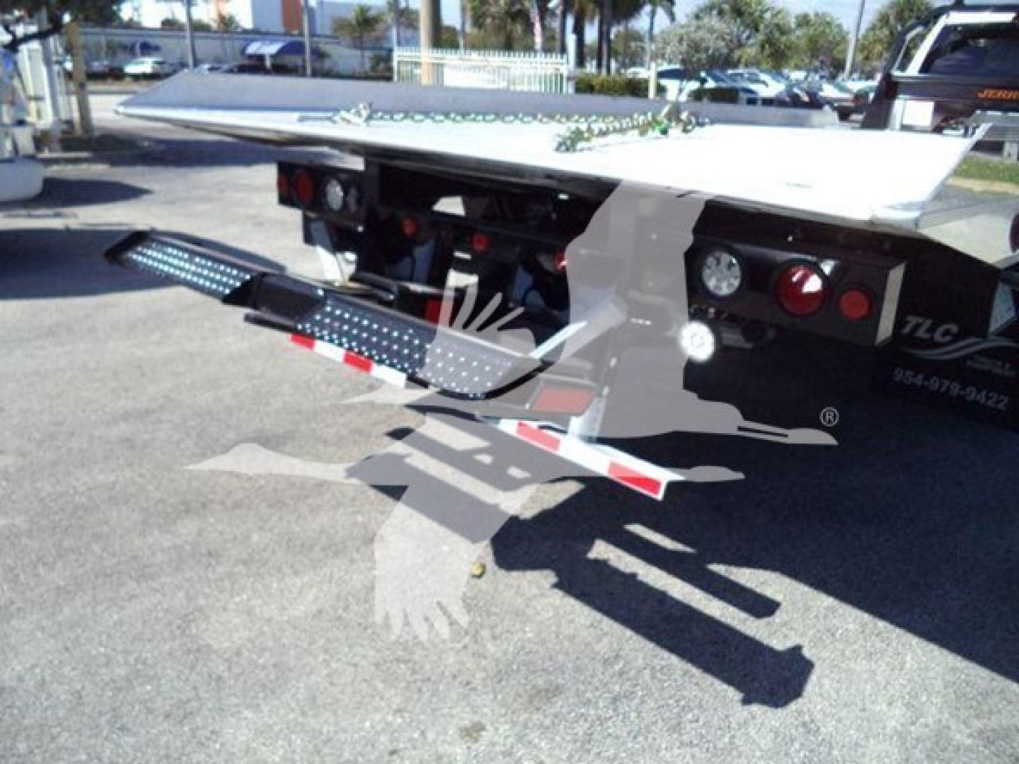Equipment trailer with hydraulic rollback bed and steps extended, Ram 1500 pickup in distance; rear of rollback trailer showing platform, taillights, and safety reflectors.