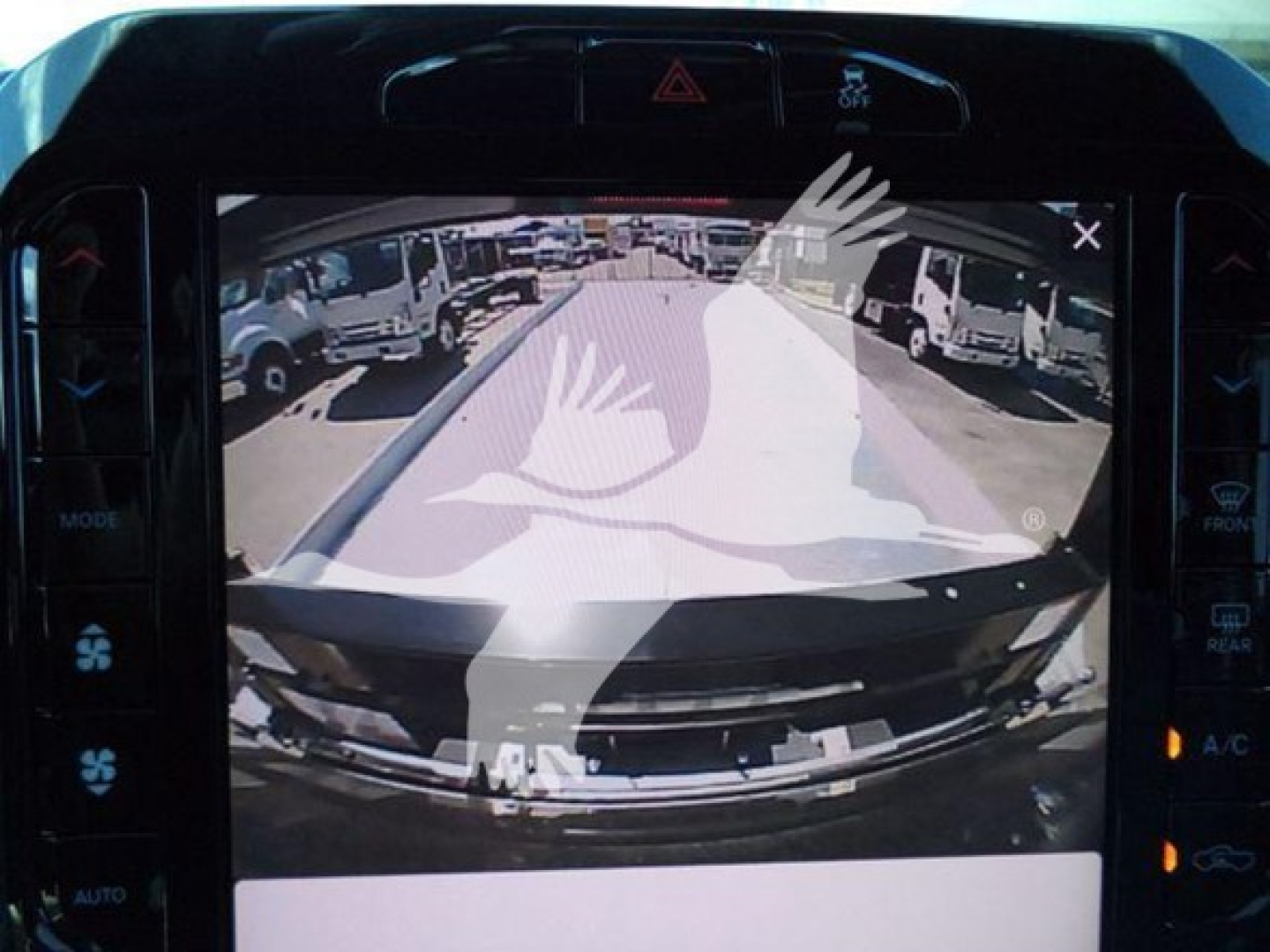 Vehicle interior display showing a rear-view camera feed of a Ram truck with a large white watermark across the image, dashboard controls visible.