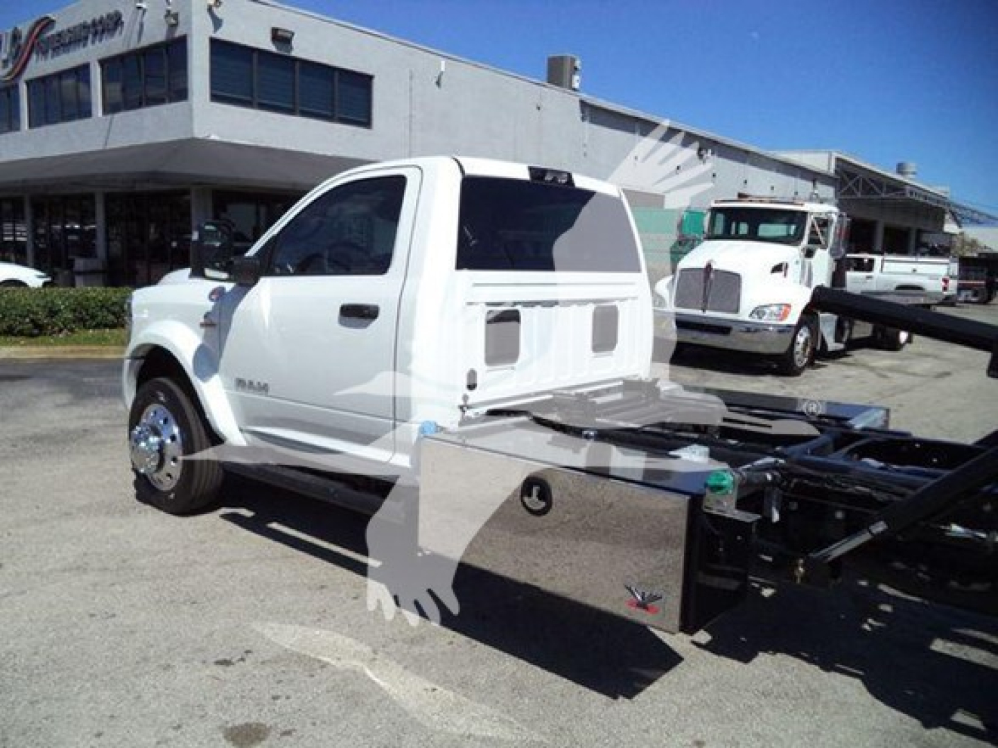 Ram pickup truck in a dealership lot with rollback chassis and flat-bed equipment visible, white cab, black rims, showroom building in background.