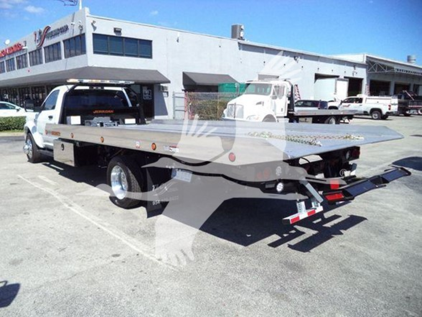 Flatbed tow truck in a dealership lot with white Ram pickup cab, flatbed equipment, and loading ramps visible.