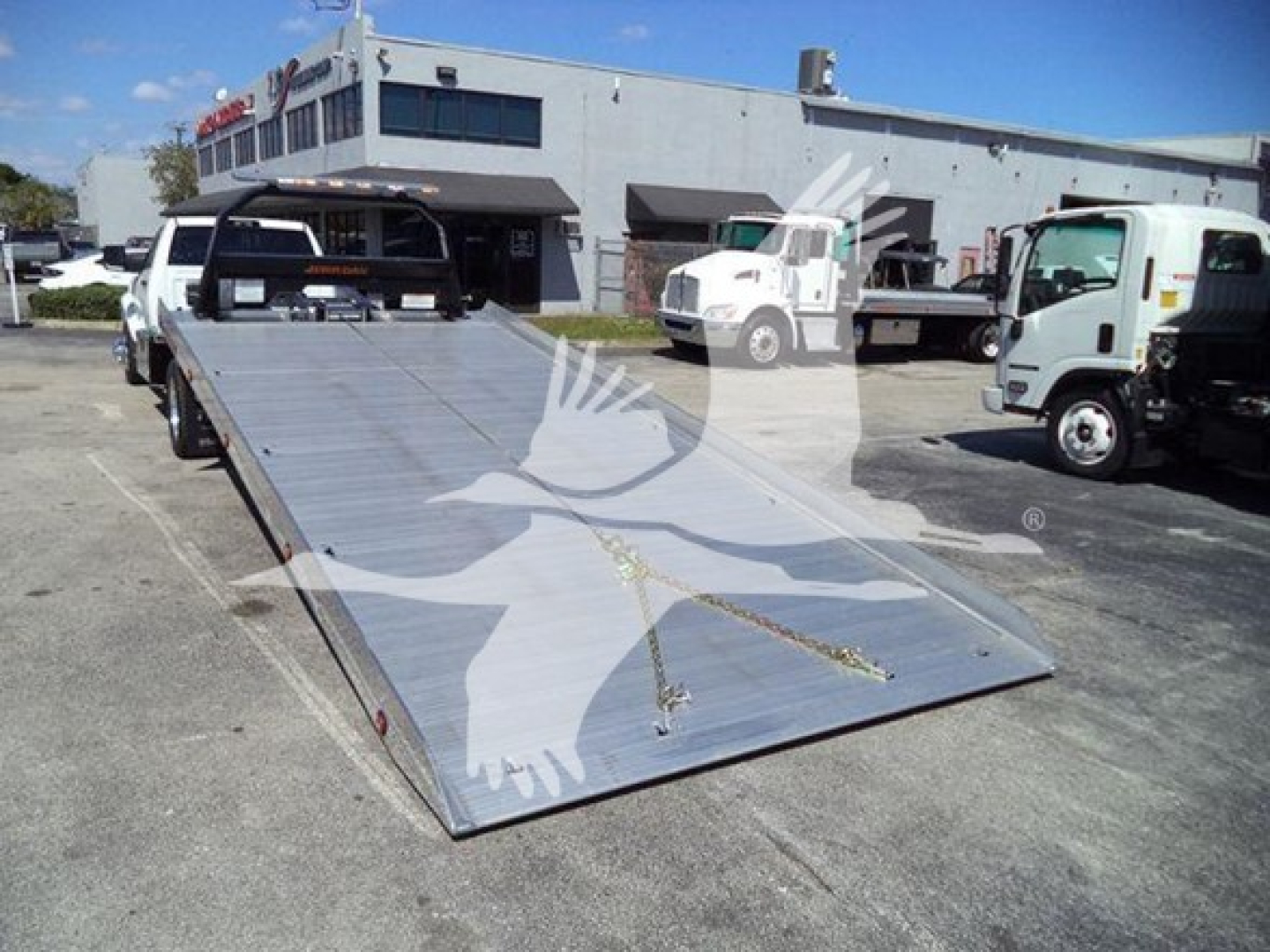 Flatbed tow truck bed with Ram logo overlay; aluminum surface with chain and hook, parked in lot with other trucks.