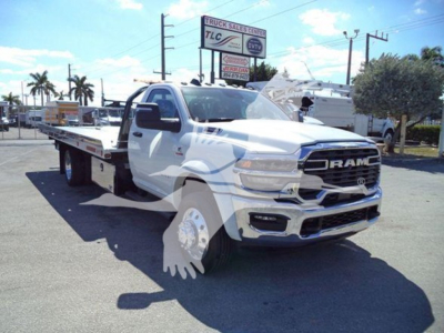 White Ram tow truck/rollback with extended flatbed and large chrome grille showing RAM badge and front bumper. Vehicle in dealership lot with palm trees and sign in background.