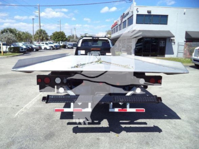 Flatbed rollback tow truck in a dealership lot, rear view showing metal platform and tail lights, with building and other vehicles in background.