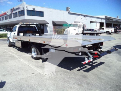Flatbed tow truck in a dealership lot with white Ram pickup cab, flatbed equipment, and loading ramps visible.