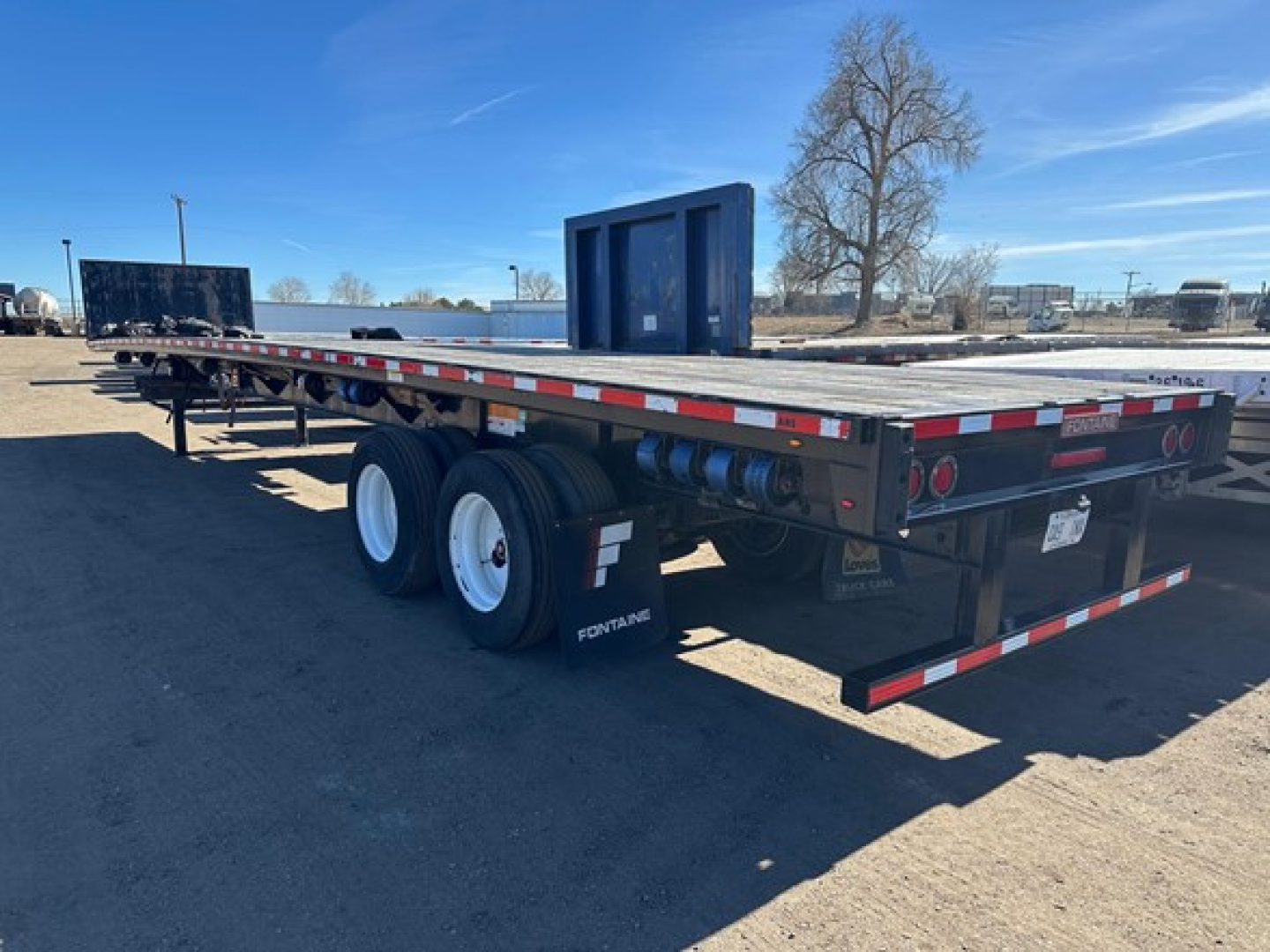 Flatbed trailer with dual rear axles and black steel frame showing Fontaine branding on mudflaps and wheel hubs; rear bumper with red/white reflective tape; blue cab-over wheel ramp in background.