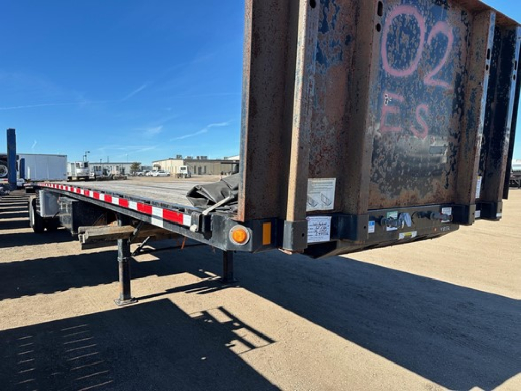 Flatbed trailer rear corner showing steel framework with ramp and support legs, orange reflectors, and weathered surface.