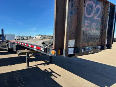 Flatbed trailer rear corner showing steel framework with ramp and support legs, orange reflectors, and weathered surface.